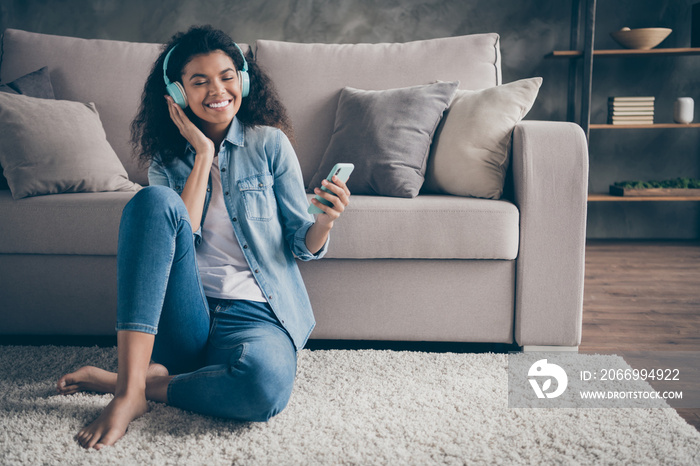 Photo of beautiful dark skin wavy lady closed eyes holding telephone wear cool earflaps listening new audio sitting floor near couch casual denim outfit living room indoors