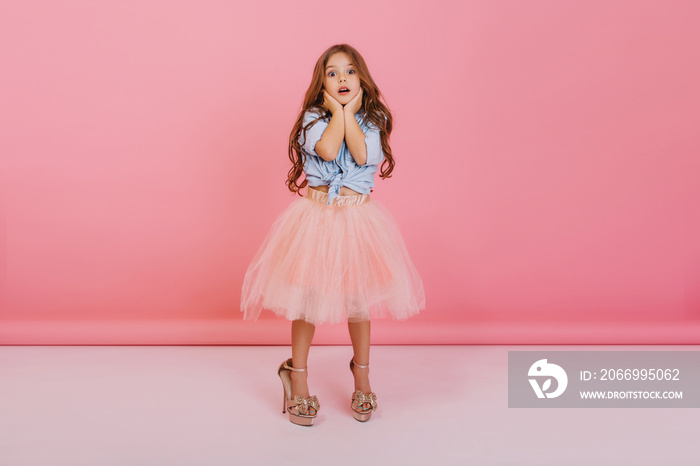 Lovely moments of cute little astonished girl expressing to camera isolated on pink background. Wearing mothers shoes, fashionable young child in tulle skirt, blue jeans shirt