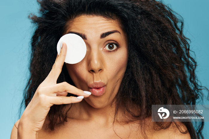 Closeup photo of excited pretty naked young Latin curly woman posing with cotton pads, covering her eye and looking at camera, using makeup removing beauty products, isolated blue background