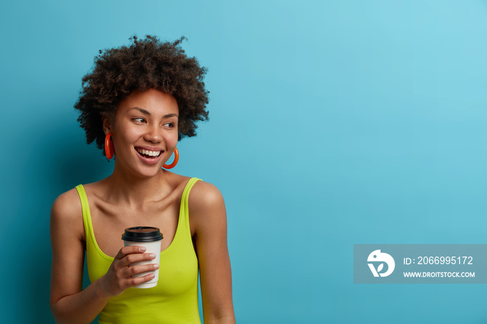 Portrait of cheerful young Afro American woman holds paper cup of coffee or espresso, smiles broadly, dressed in casual vest, enjoys spare time, concentrated aside, isolated on blue background