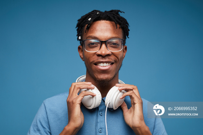 Head and shoulders portrait of contemporary African-American man wearing headphones smiling at camera while posing against blue background, copy space