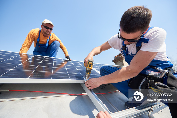 Male team workers installing solar photovoltaic panel system using screwdriver. Two electricians mounting blue solar module on roof of modern house. Alternative energy innovation concept.