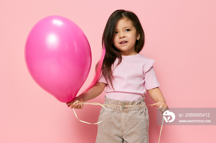 cheerful, cute, playful preschool age girl playing with a pink balloon in her hand smiling joyfully at the camera standing on a pink background with empty space for advertising mockup insert