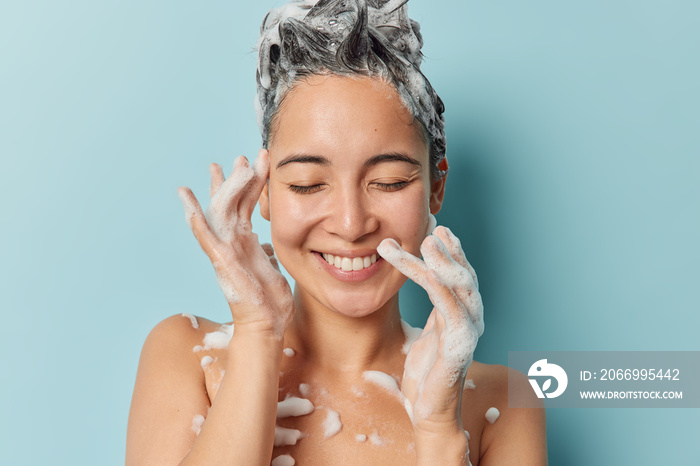 Pleased young brunette Asian woman washes hair applies shampoo smiles gently keeps eyes closed stands naked has soap foam on body healthy smooth skin takes shower isolated over blue background