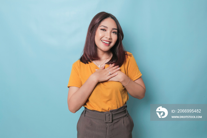 Happy mindful thankful young woman holding hands on chest smiling isolated on blue background feeling no stress, gratitude, mental health balance, peace of mind concept.