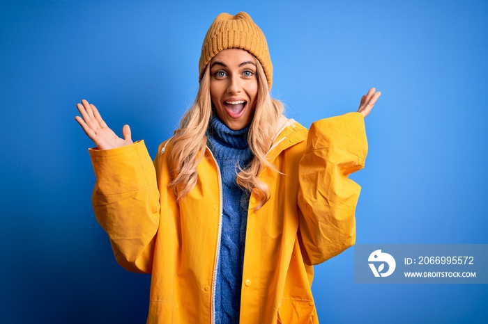 Young beautiful blonde woman wearing raincoat for rainy weather over blue background celebrating crazy and amazed for success with arms raised and open eyes screaming excited. Winner concept