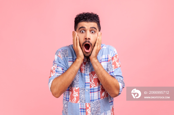 Portrait of astonished bearded man in blue shirt looking with big eyes, holding hands on face and screaming in amazement, shocked by sudden event. Indoor studio shot isolated on pink background.