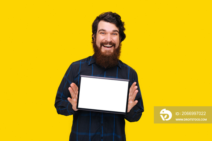 Cheerful bearded guy holding a tablet, and showing white screen blank, over yellow background