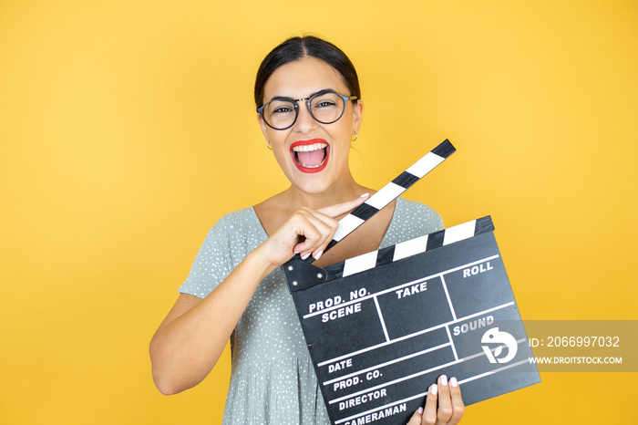 Young beautiful woman wearing glasses standing over isolated yellow background holding clapperboard very happy
