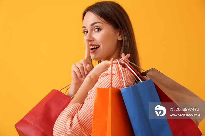 Beautiful young woman with shopping bags showing silence gesture on color background