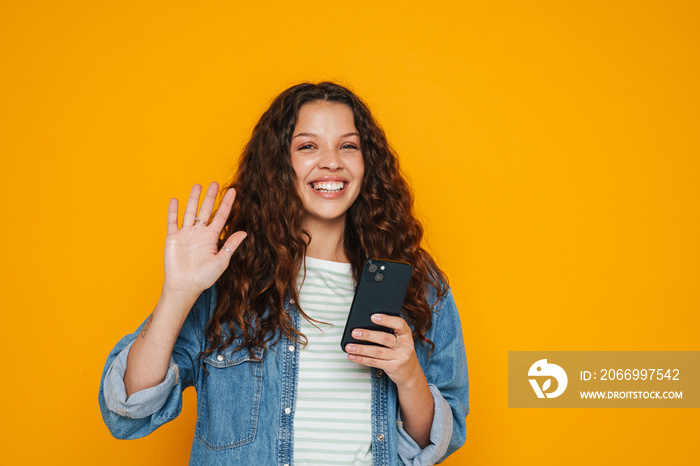 Excited girl holding cellphone and waving isolated over yellow background