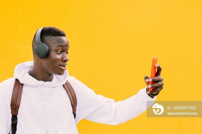 Young african American black man looking at smart phone while listening music with wireless headphones. Taking self portrait, selfie. Yellow wall wearing a white sweatshirt and a backpack