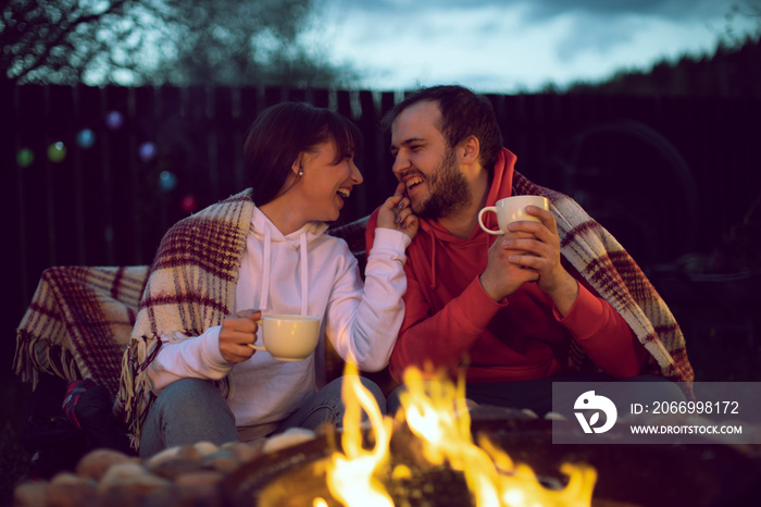 Happy Lovers communicate and laugh by the fire. Married couple is relaxing and drinking tea in nature. Rest in the country with a camper during quarantine.