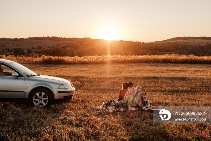 Happy Young Family Mom and Dad with Their Little Son Enjoying Summer Weekend Picnic on the Car Outside the City in the Field at Sunny Day Sunset, Vacation and Road Trip Concept