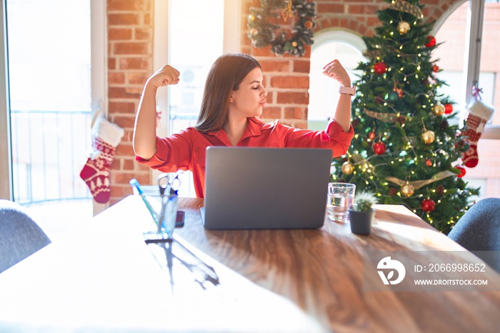 Beautiful woman sitting at the table working with laptop at home around christmas tree showing arms muscles smiling proud. Fitness concept.