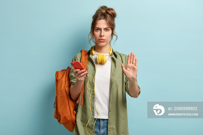 Serious cute woman spends time indoor with modern gadget, shows stop gesture, asks to slow down, travels with music and rucksack, dressed in sylish outfit, makes denial sign, isolated on blue