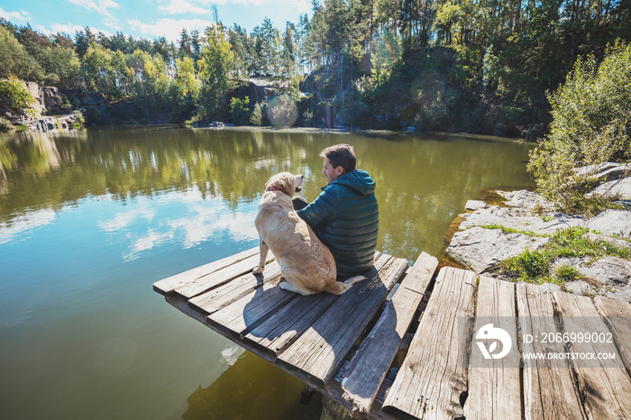 A man with a Labrador retriever dog sits on a wooden deck on a beautiful rocky shore of a lake and looks at the water