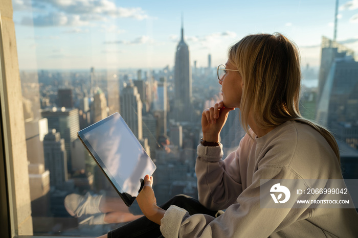 Thoughtful female digital nomad with modern touch pad looking in panoramic window while creation social content for sharing to web blog, millennial hipster girl with gadget for online communication