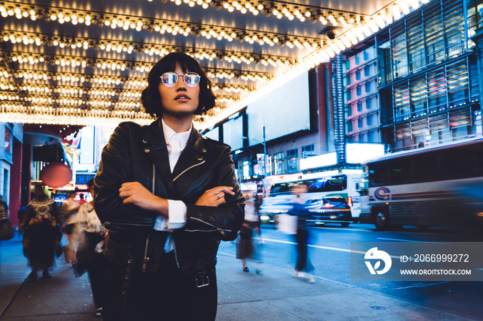 Thoughtful female tourist in optical spectacles with neon reflection thinking about travel way for exploring night New York, pondering hipster girl hanging out to downtown for evening resting