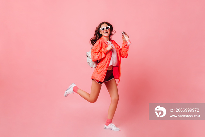 Sports girl in windbreaker, shorts and sneakers runs on pink background. Woman in sunglasses holding bottle of water