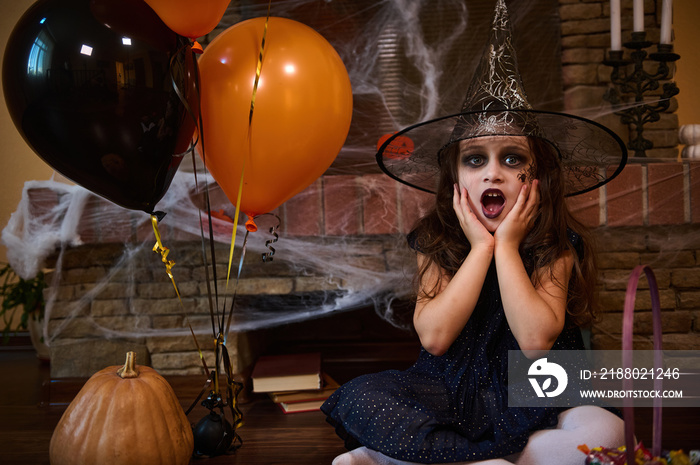 Charming Caucasian little girl, dressed in witch and wizard hat, sitting near orange black balloons 