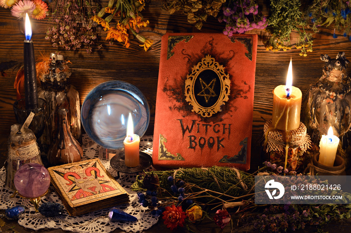 Still life with tarot cards, crystal ball, candles and witch book with hanging herbs on table.