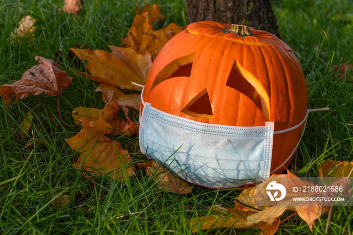 Carved Halloween pumpkin with sad eyes wearing mask on the grass with leaves, no party during Covid 