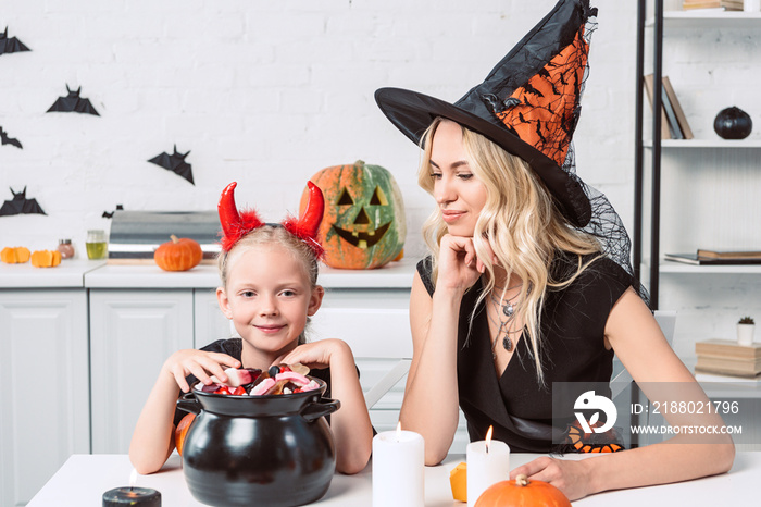 mother and little daughter in halloween costumes at table with treats in black pot in kitchen