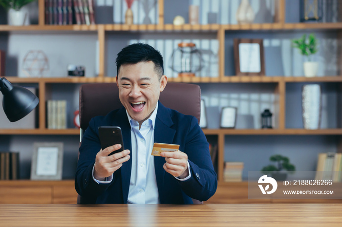 Young handsome asian man in suit in the office at work holding a credit card and shopping, ordering 