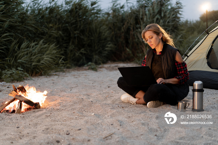 Evening tent camping in seashore. Female traveler uses laptop computer sitting by campfire. Woman on