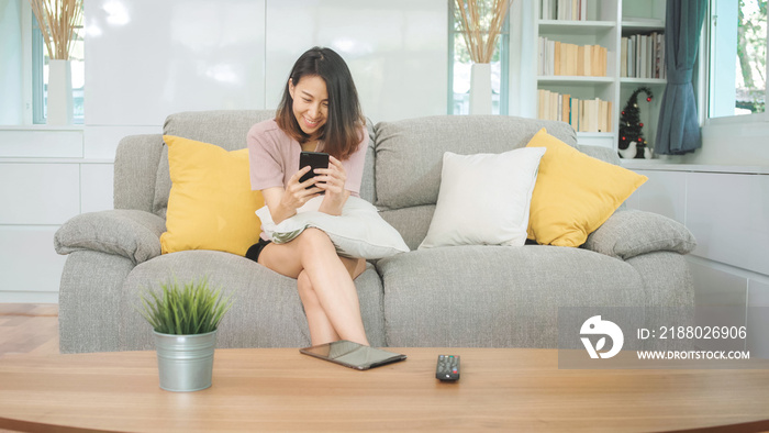 Young Asian woman using smartphone checking social media feeling happy smiling while lying on the so