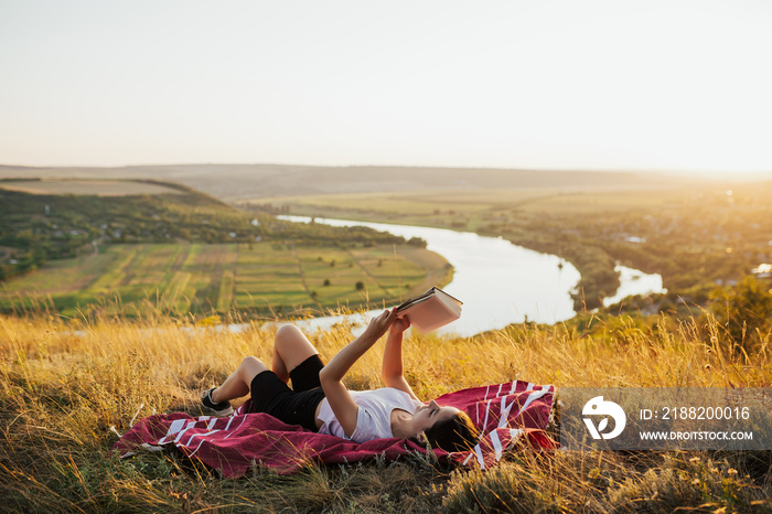 Young beautiful woman lying on a  blanket on the grass on the hill and reading a book. She read a book with beautiful landscape on background. Copy space.