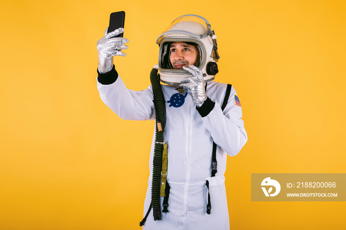 Male cosmonaut in space suit and helmet, taking a selfie with mobile phone, on yellow background.