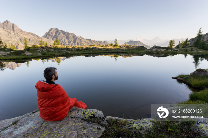 Young adult man in red sleeping bag near mountain lake with reflection at dawn in summer outdoor.
