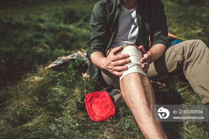 First medical care in travelling. Close up portrait of young traveler male with bandaged leg sitting on green grass hill. Copy space on left