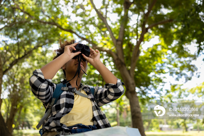 Young women teenager ethnic African American black skin wearing plaid shirt and her backpack sitting at tree base looking binocular in her hand