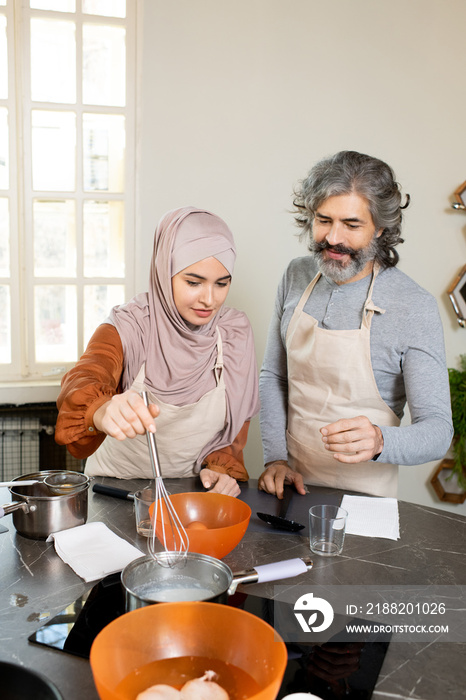 Young Muslim female trainee in hijab holding whisker over metallic pan on stove