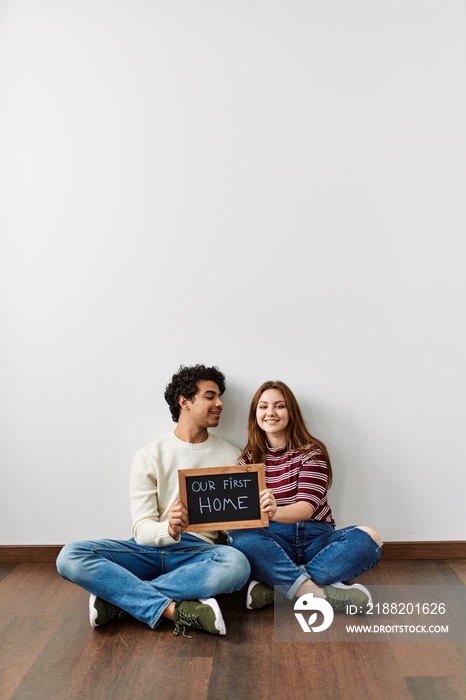 Young hispanic couple holding our first home blackboard sitting on the floor at empty new house.