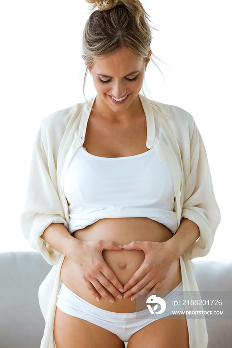 Young beautiful pregnant woman looking belly with heart made up of fingers at home.