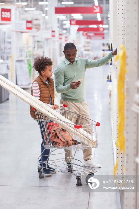 Vertical full length portrait of African-American father and son shopping together in hardware store and using smartphone while choosing wooden boards for home improvement.