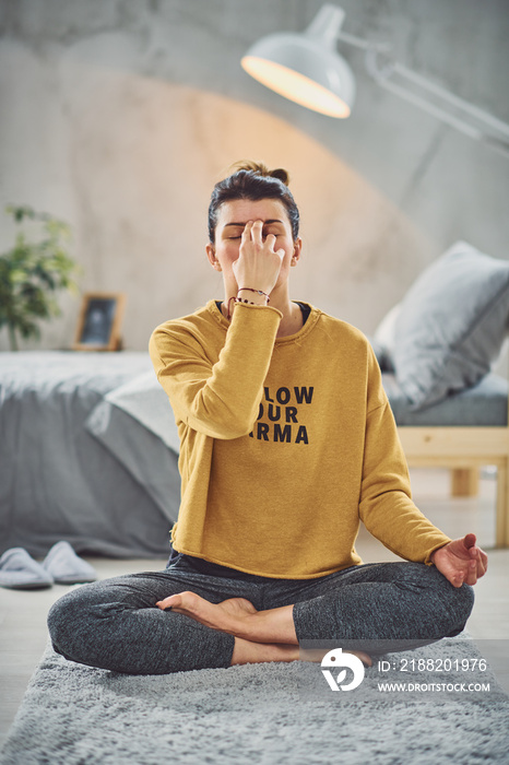 Beautiful Caucasian brunette sitting in lotus yoga pose and practicing breathing. Bedroom interior, morning routine.