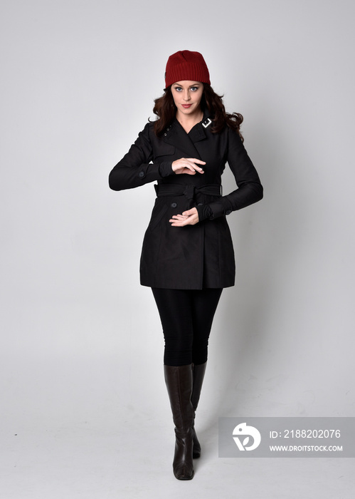 full length portrait of a pretty brunette girl wearing a black coat, leather boots and red beanie. Standing pose  on a grey studio background.