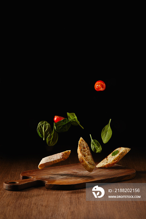 Bread pieces and salad leaves with tomatoes flying above wooden table surface