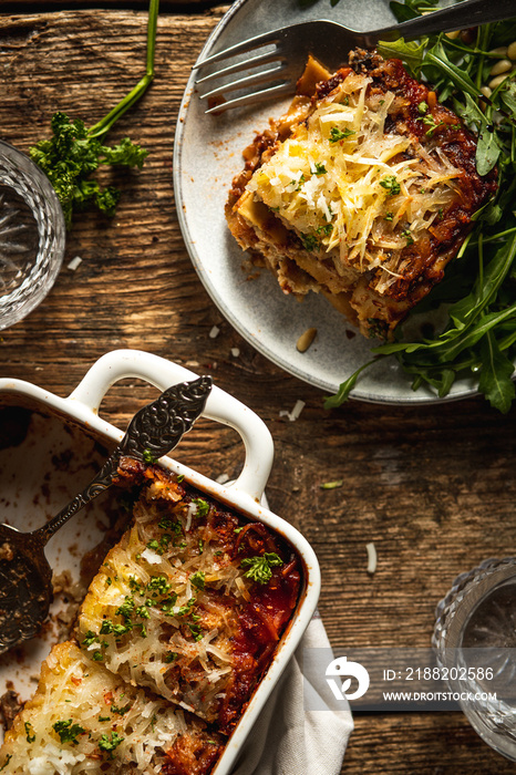 Baked Vegan lasagna casserole with roasted eggplant and mushrooms on a rustic wooden table served on a plate with some arugula  and a lot of vegan shredded cheese and fresh herbs on top