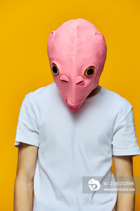 a man in a funny rubber mask looks at the floor while standing on a yellow background