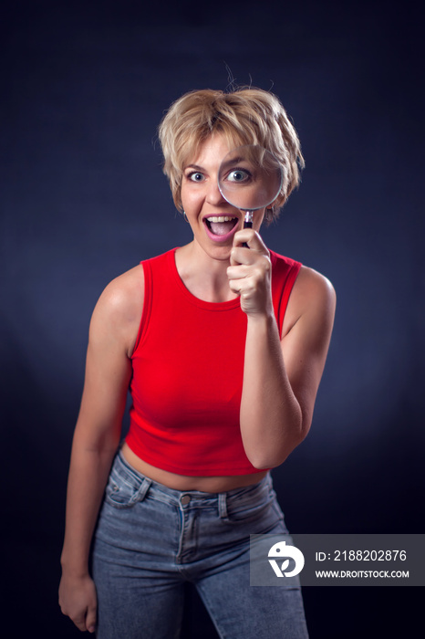 A portrait of young woman with short blond hair in red shirt holding magnifier in front of eye beside of black background. People and emotions concept