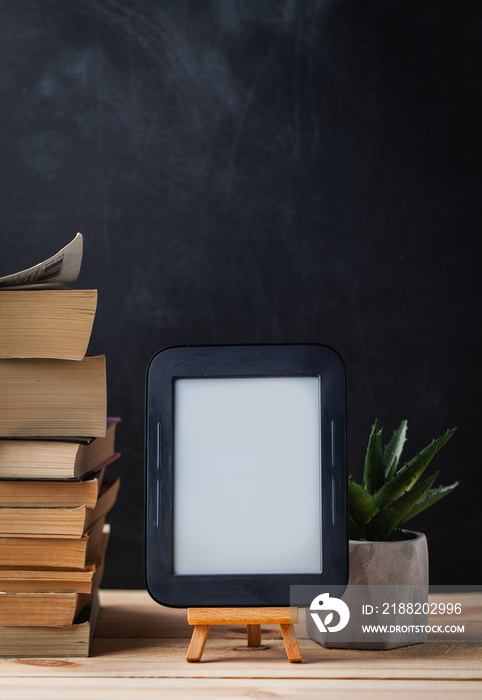 E-book reader on the wooden stand next to the stack of paper books at the black background.