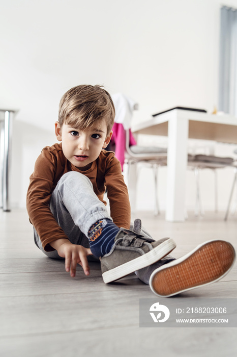 Full length view of small cute caucasian boy little child kid sitting on the wooden or laminated vinyl floor at home putting on or taking off the shoes in day alone front view