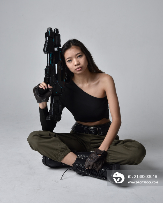 Full length portrait of pretty brunette, asian girl wearing black top and khaki utilitarian army pants and leather boots. Sitting pose holding a science fiction gun, isolated agent a light grey studio