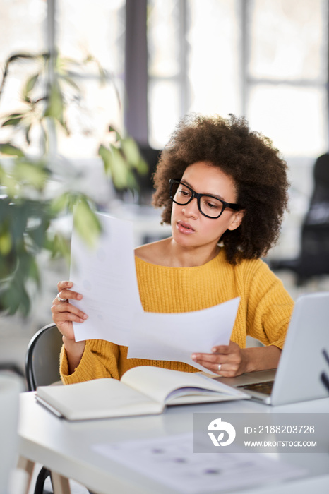 Serious mixed race attractive businesswoman dressed smart casual holding paperwork while sitting in modern office.